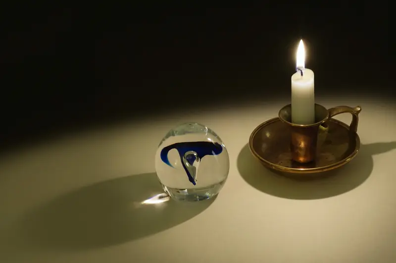 Lit candle in a brass holder beside a glass sphere on a cream background