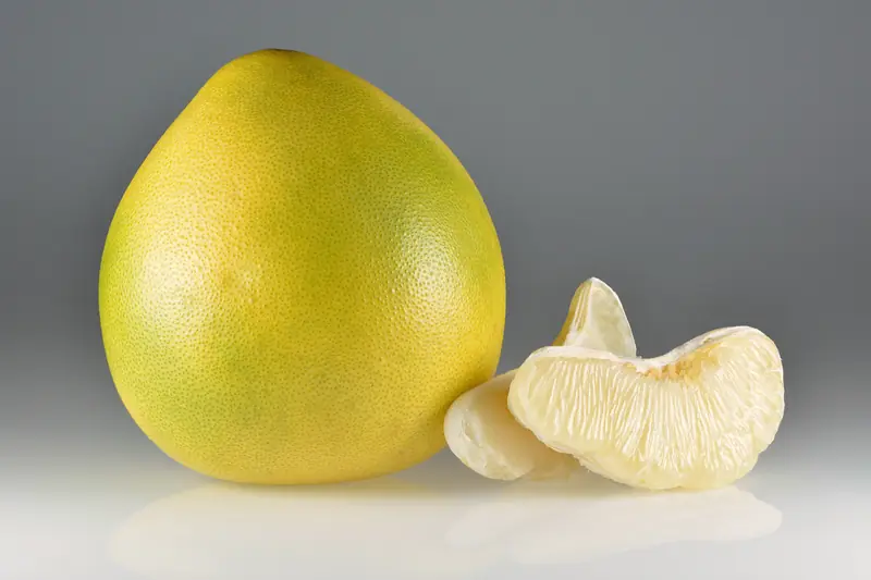 Pomelo vert-jaune entier posé à côté de quartiers pelés à la chair pâle
