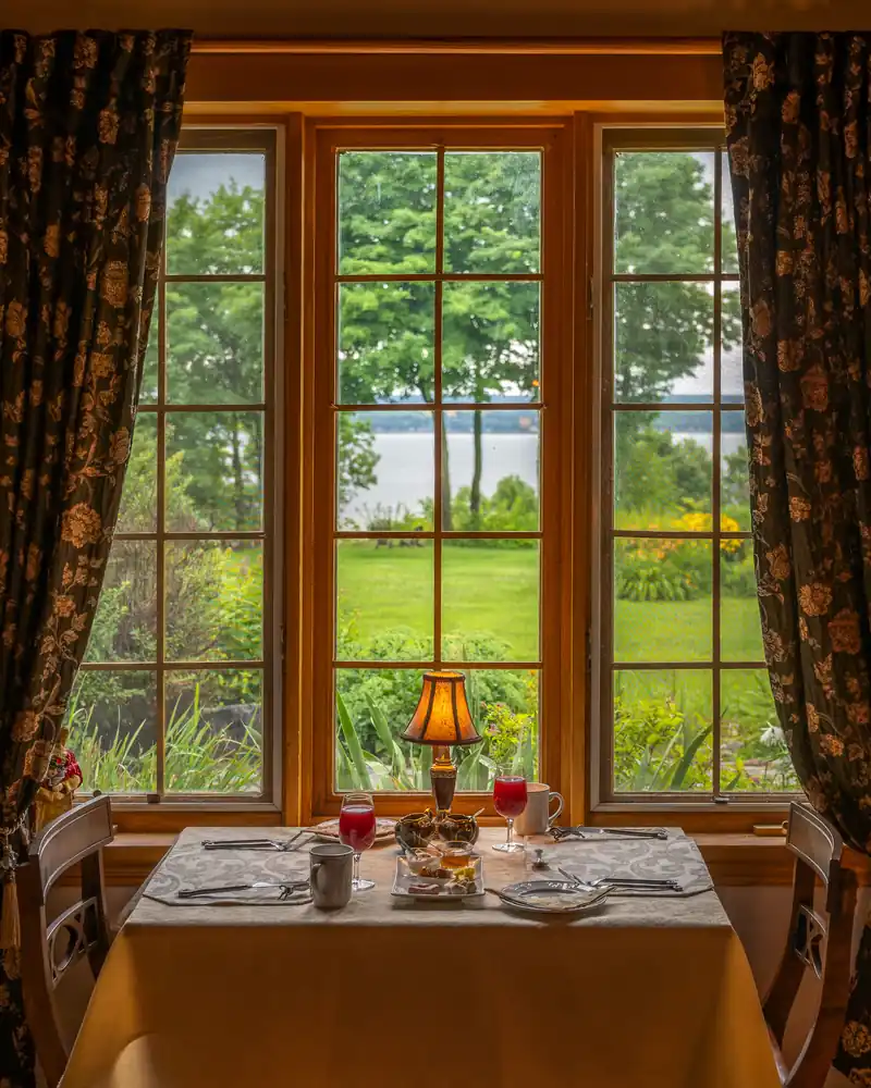 Table de petit-déjeuner dressée devant une grande fenêtre ouverte sur un jardin et un fleuve
