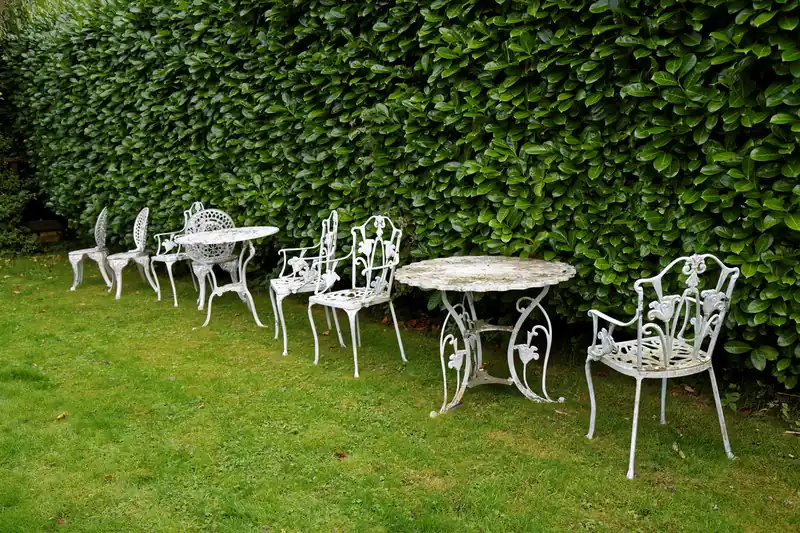 Tables et chaises de jardin en fonte blanche alignées sur l'herbe contre une haie