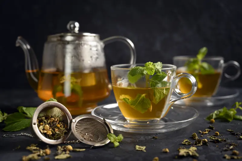 Glass teapot and cups of mint tea on a dark table, with an open tea infuser
